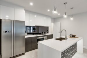 CANBERRA, AUSTRALIA – JUNE 2, 2018: New kitchen furnished with stainless steel fridge, reflective glass splashback, island, inset sink, stove, oven and three hanging lights in a modern home